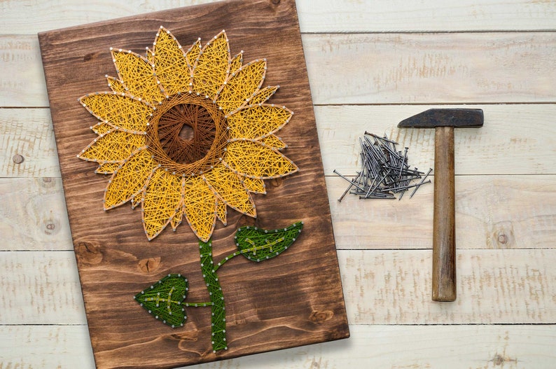 Pu&ograve; includere: Un pannello di legno con un design di girasole in arte con la corda. Il girasole &egrave; realizzato con corda gialla e marrone e ha foglie verdi. Sono mostrati anche un martello e chiodi.