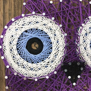 May include: A handcrafted string art owl design. The owl features large, detailed eyes with concentric circles of white, light blue, dark blue, and black. The owl's body and the background are made of purple string, all mounted on a wooden board.