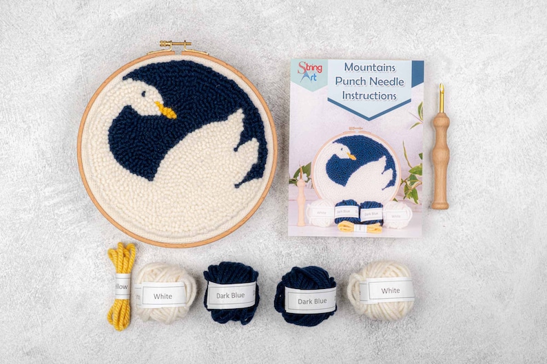 K&ouml;nnte beinhalten: Punch-Needle-Set f&uuml;r ein Schwanendesign. Das Set enth&auml;lt einen Holzreifen, Garn in Wei&szlig; und Dunkelblau, eine Punch-Needle und eine Anleitung. "Mountains Punch Needle Instructions" ist auf dem Anleitungsheft gedruckt.