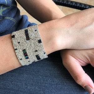 以下が含まれることがあります： 幾何学模様の銀と黒のビーズブレスレット。ブレスレットは手首につけられています。