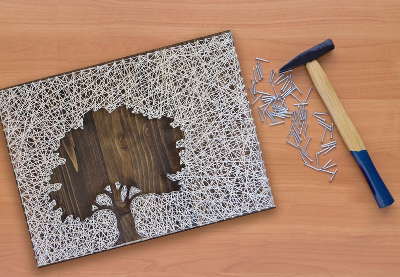 Puede incluir: Una pieza de arte de cuerda blanca en una tabla de madera que representa un &aacute;rbol. El arte de cuerda est&aacute; hecho con clavos y cuerda. Tambi&eacute;n hay un martillo y clavos en la imagen.