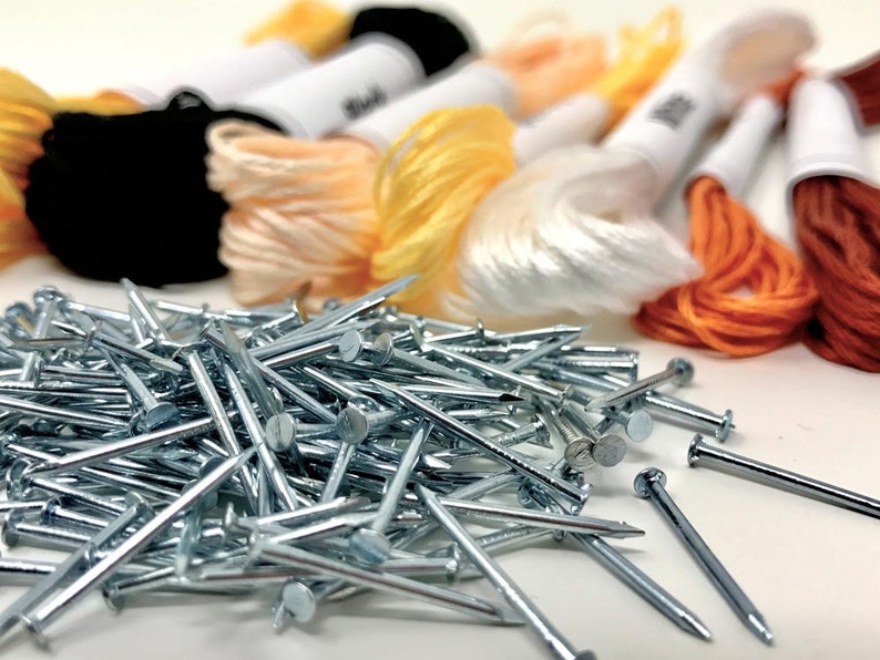 Puede incluir: Primer plano de un mont&oacute;n de tachuelas o clavos de color plateado. Las tachuelas est&aacute;n dispuestas en un patr&oacute;n aleatorio y est&aacute;n rodeadas de carretes de hilo de bordar de colores en tonos naranja, amarillo y blanco.