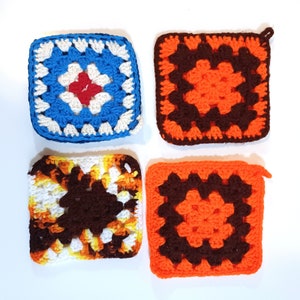 May include: Four square crochet potholders in various colours. One is blue with white and red, another is orange with brown, a third is white, brown, and yellow, and the last is orange with brown. Each has a distinct pattern.