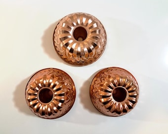 Puede incluir: Tres moldes para pasteles de color cobre con bordes acanalados. Los moldes son de diferentes tamaños y están dispuestos en forma triangular.