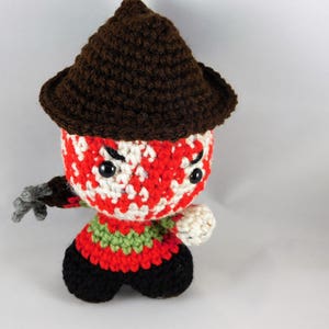 Pode incluir: Um brinquedo de pel&uacute;cia de croch&ecirc; de um personagem de filme de terror usando um chap&eacute;u marrom. O brinquedo tem listras vermelhas, brancas e verdes e olhos pretos.