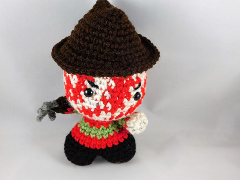 Pode incluir: Um brinquedo de pel&uacute;cia de croch&ecirc; de um personagem de filme de terror usando um chap&eacute;u marrom. O brinquedo tem listras vermelhas, brancas e verdes e olhos pretos.