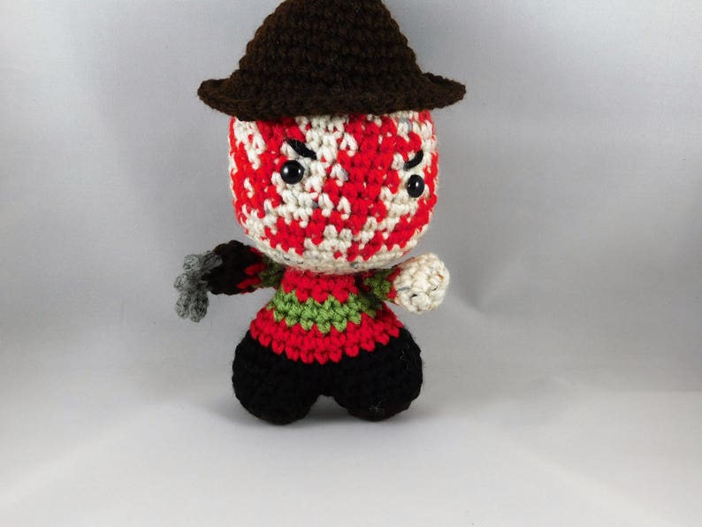 Pode incluir: Um brinquedo de pel&uacute;cia de croch&ecirc; de um personagem de filme de terror. O brinquedo est&aacute; usando um chap&eacute;u marrom e tem um rosto listrado de vermelho e branco com olhos pretos. O brinquedo est&aacute; segurando uma faca na m&atilde;o direita.