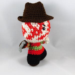 Pode incluir: Um brinquedo de pel&uacute;cia de croch&ecirc; de um personagem de filme de terror usando um chap&eacute;u marrom e segurando uma faca. O personagem tem uma m&aacute;scara listrada de vermelho e branco.