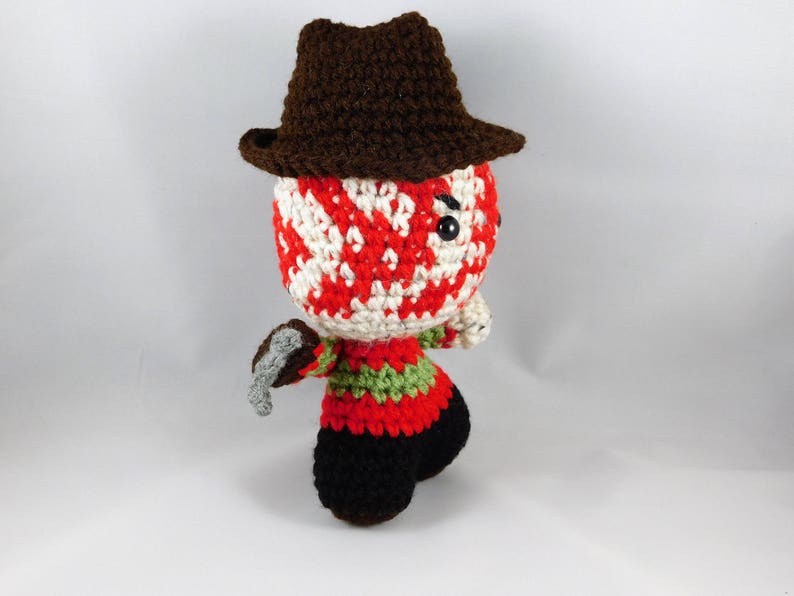Pode incluir: Um brinquedo de pel&uacute;cia de croch&ecirc; de um personagem de filme de terror usando um chap&eacute;u marrom e segurando uma faca. O personagem tem uma m&aacute;scara listrada de vermelho e branco.