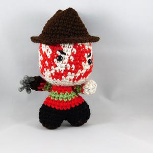 Pode incluir: Um boneco de Freddy Krueger de croch&ecirc; com um chap&eacute;u marrom, um rosto listrado de vermelho e branco e um su&eacute;ter listrado de verde e vermelho. O boneco est&aacute; segurando uma garra de prata.