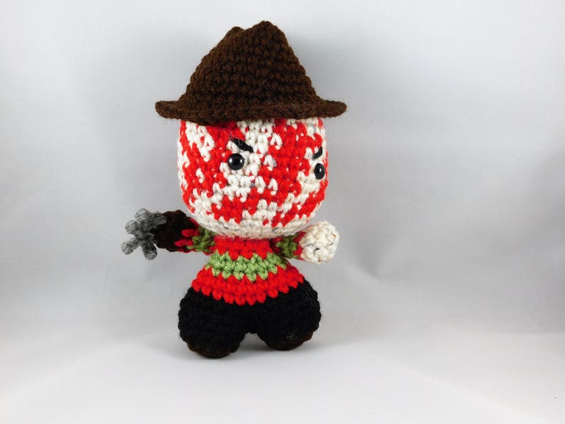 Pode incluir: Um boneco de Freddy Krueger de croch&ecirc; com um chap&eacute;u marrom, um rosto listrado de vermelho e branco e um su&eacute;ter listrado de verde e vermelho. O boneco est&aacute; segurando uma garra de prata.