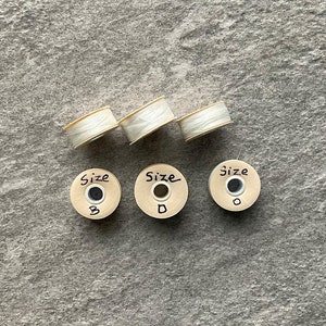May include: Four white spools of thread with labels that say "Size B", "Size D", and "Size O".