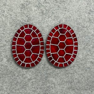 May include: Two oval-shaped cabochons featuring a deep red, glossy surface divided by a silver-toned grid pattern that resembles a turtle shell. Each piece has a smooth, rounded edge and a consistent geometric honeycomb design across the front.