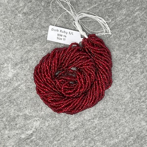 May include: A close-up of a spool of dark ruby red seed beads. The beads are size 11/0 and are arranged in a circular pattern. The label reads "Dark Ruby S/L NW-14 Size 11".