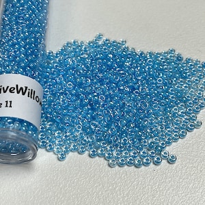 May include: A close-up of a small clear container filled with light blue seed beads. The container has a white label that reads "NativeWillow Size 11". A pile of the same light blue seed beads is scattered on a white surface.
