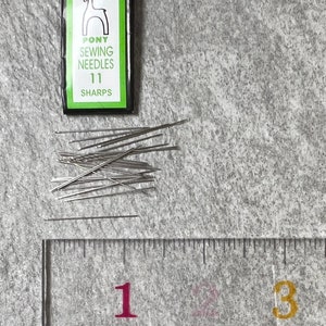May include: A package of Pony Sewing Needles 11 Sharps, with a white horse logo on a green and black label. Several silver sewing needles are scattered beside a clear ruler, with inch markings.