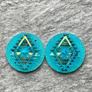 May include: Two turquoise glittery round earrings with a geometric diamond design in the center. The diamond has yellow and blue accents. The earrings are a bright, eye-catching accessory.