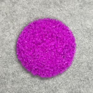 May include: A circular container filled with vibrant magenta seed beads. The beads are small and uniform in size, creating a dense, textured surface. The container is transparent, and the background is a light gray, textured surface.