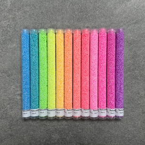 May include: A collection of colorful craft beads in clear plastic tubes. The tubes are arranged in a row, showcasing a rainbow of colors including blue, green, yellow, orange, pink, and purple. Each tube is labeled with white text.