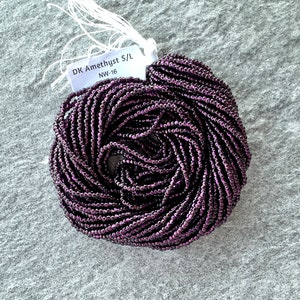May include: A close-up of a spool of dark amethyst seed beads. The beads are small and round, with a metallic sheen. The spool is labeled "DK Amethyst S/L NW-16".