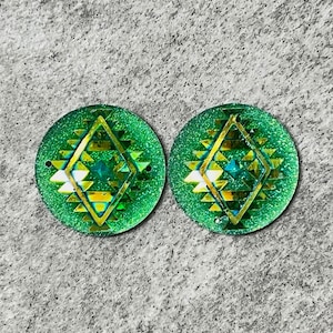 May include: Two green round resin cabochons with a gold and white geometric design. The cabochons have a sparkly, glittery finish.