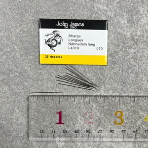 May include: A yellow and black packet of 25 John James Sharps Longues sewing needles, size 010, with the text "L4310" and "25 Needles" printed on the front. The needles are laid out on a white ruler with black markings.
