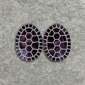 May include: Two oval-shaped earrings with a dark purple, mosaic-like design. Each earring has a silver-colored border and a pattern of white lines creating a honeycomb effect. The earrings are set against a gray background.