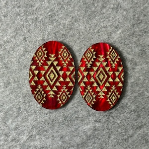 May include: Two oval-shaped earrings with a red background and a gold geometric pattern. The earrings have a tribal design with diamond and triangle shapes. The earrings are displayed on a gray surface.