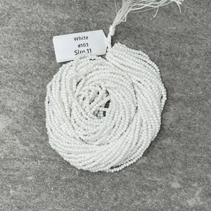 May include: A bundle of white seed beads, size 11, arranged in a circular pattern. A white tag attached to the beads reads "White #103 Size 11". The beads are on a gray surface.