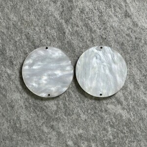 May include: Two white round acrylic blanks with a swirl pattern. The blanks have a hole in the top for hanging.