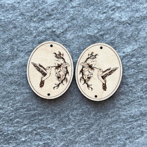 May include: Two oval-shaped wooden pendants with a hummingbird and floral design. The hummingbird is depicted in flight, with its beak near a cluster of flowers. The pendants have small holes at the top and bottom for stringing.