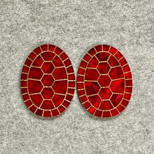May include: A pair of oval earrings with a red and gold design. The earrings have a mosaic pattern with a red centre and a gold border. The centre is divided into hexagonal shapes. The earrings are on a grey background.