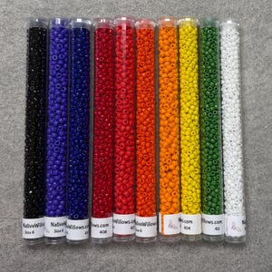 May include: Ten clear plastic tubes filled with small, round, opaque seed beads in a spectrum of colors including black, purple, dark blue, red, orange, yellow, green, and white. Each tube features a white label at the base with text.