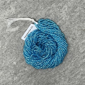 May include: A close-up of a spool of aquamarine blue seed beads. The beads are small and square-shaped, and they are arranged in a tight coil. The spool is labeled "Aquamarine 5/0".