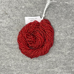 May include: A strand of red seed beads, labelled "Lt Ruby S/L NW-151", is coiled into a circular shape. The beads are small and uniform in size, creating a smooth, textured surface.