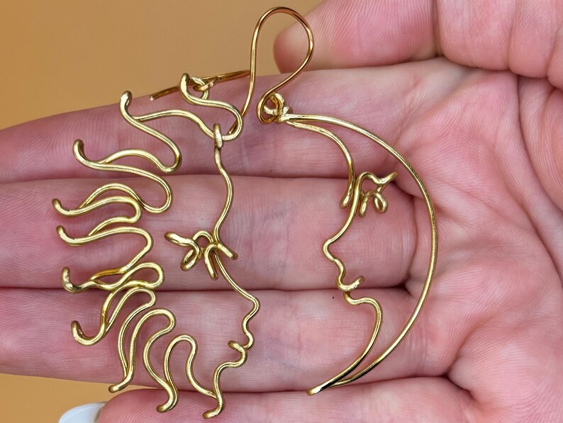 Détails rapprochés des boucles d’oreilles en forme de soleil et de lune, confectionnées à la main en fil doré. Design élégant et minimaliste inspiré du cosmos.