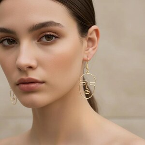 Femme portant des boucles d’oreilles visage abstrait en fil doré, design minimaliste fait main. Style élégant et artistique, lignes continues formant un visage. Photo lifestyle avec lumière naturelle douce, fond beige neutre, esthétique raffinée type boutique parisienne.