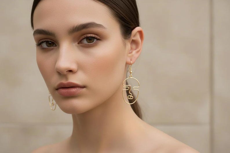 Femme portant des boucles d’oreilles visage abstrait en fil doré, design minimaliste fait main. Style élégant et artistique, lignes continues formant un visage. Photo lifestyle avec lumière naturelle douce, fond beige neutre, esthétique raffinée type boutique parisienne.