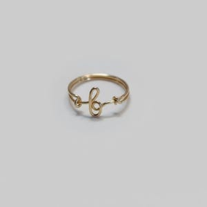 May include: A gold-coloured ring with a delicate design. The ring features a cursive letter "b" in the centre, with a simple band. The ring is likely made of gold-toned metal and is set against a white background.