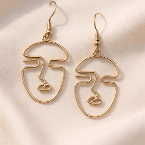 Boucles d’oreilles visage abstrait en fil doré, style minimaliste fait main. Design artistique avec lignes continues formant un visage, finition dorée douce type champagne. Présentées sur fond tissu beige clair avec lumière naturelle, rendu élégant et raffiné, style boutique parisienne.