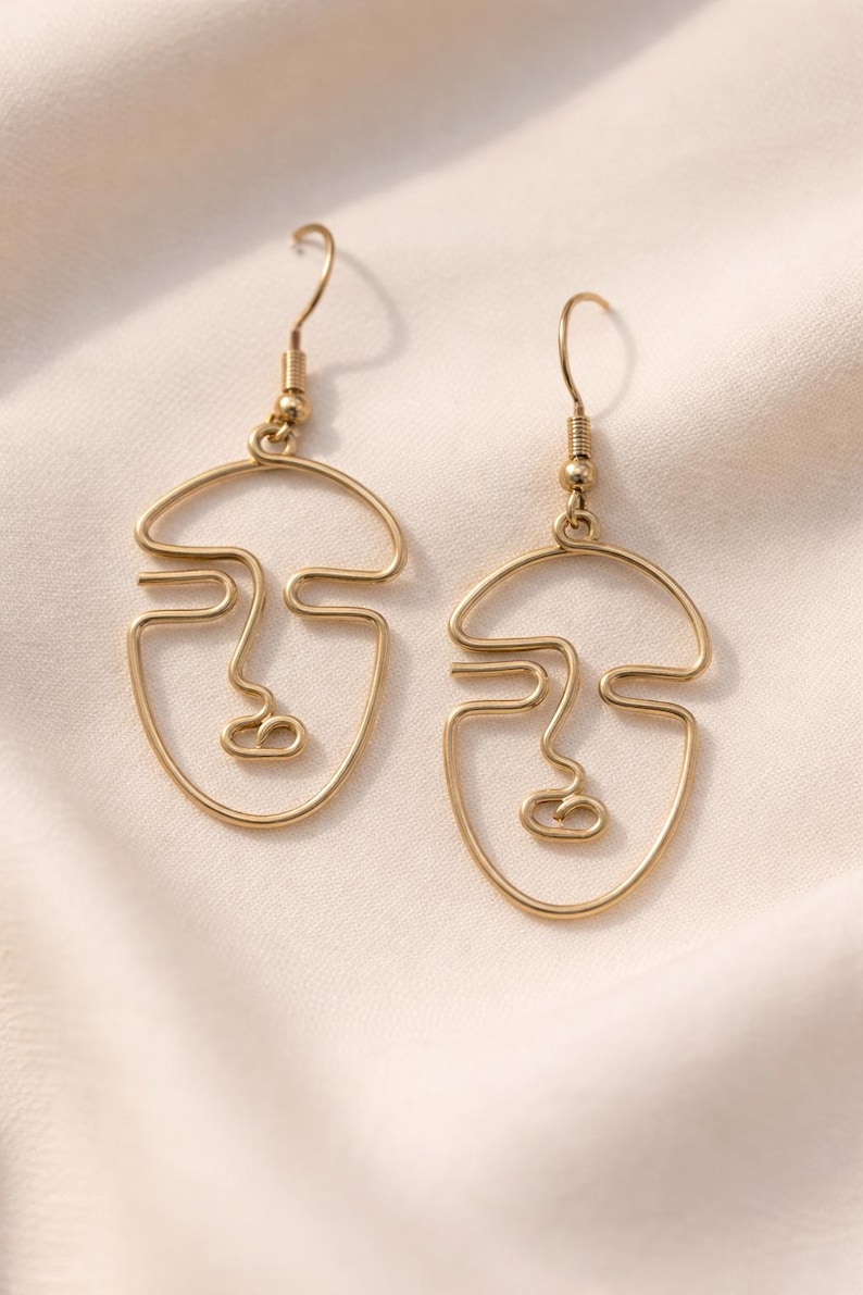 Boucles d’oreilles visage abstrait en fil doré, style minimaliste fait main. Design artistique avec lignes continues formant un visage, finition dorée douce type champagne. Présentées sur fond tissu beige clair avec lumière naturelle, rendu élégant et raffiné, style boutique parisienne.