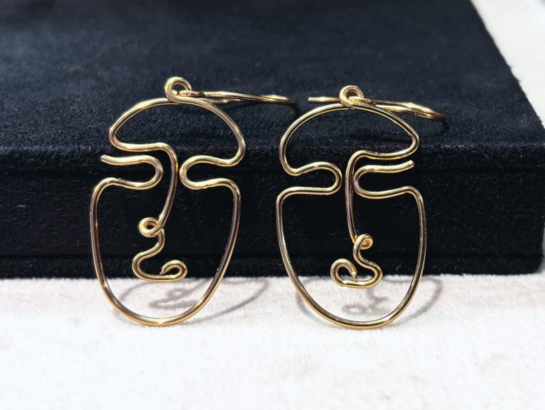 Boucles d’oreilles abstraites en fil métallique doré, représentant un visage stylisé, posées sur un fond noir et beige pour mettre en valeur leur élégance