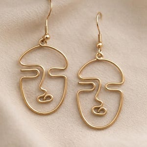 Boucles d’oreilles visage abstrait en fil doré, design minimaliste fait main. Lignes continues formant un visage artistique avec finition dorée douce. Présentées sur fond tissu beige clair avec texture légère, lumière naturelle douce et ombres subtiles, style élégant et moderne inspiré d’une boutique parisienne.