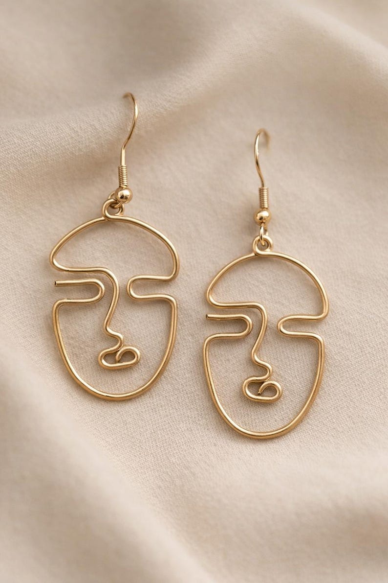 Boucles d’oreilles visage abstrait en fil doré, design minimaliste fait main. Lignes continues formant un visage artistique avec finition dorée douce. Présentées sur fond tissu beige clair avec texture légère, lumière naturelle douce et ombres subtiles, style élégant et moderne inspiré d’une boutique parisienne.