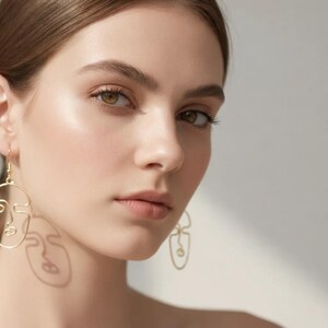 Femme portant des boucles d’oreilles visage abstrait en fil doré, design minimaliste fait main. Style artistique avec lignes continues formant un visage. Lumière naturelle douce créant une ombre légère sur le visage, fond neutre beige, rendu élégant et moderne type bijouterie parisienne.