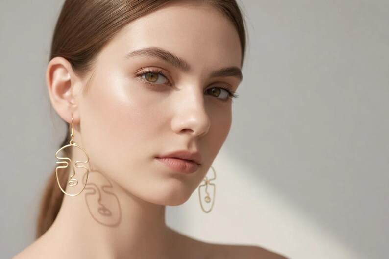Femme portant des boucles d’oreilles visage abstrait en fil doré, design minimaliste fait main. Style artistique avec lignes continues formant un visage. Lumière naturelle douce créant une ombre légère sur le visage, fond neutre beige, rendu élégant et moderne type bijouterie parisienne.