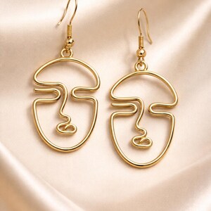 Boucles d’oreilles visage abstrait en fil doré, design minimaliste fait main. Lignes continues formant un visage artistique, finition dorée douce type champagne. Présentées sur tissu beige clair avec plis élégants, lumière naturelle douce et ombres légères, style raffiné et moderne type bijouterie parisienne.