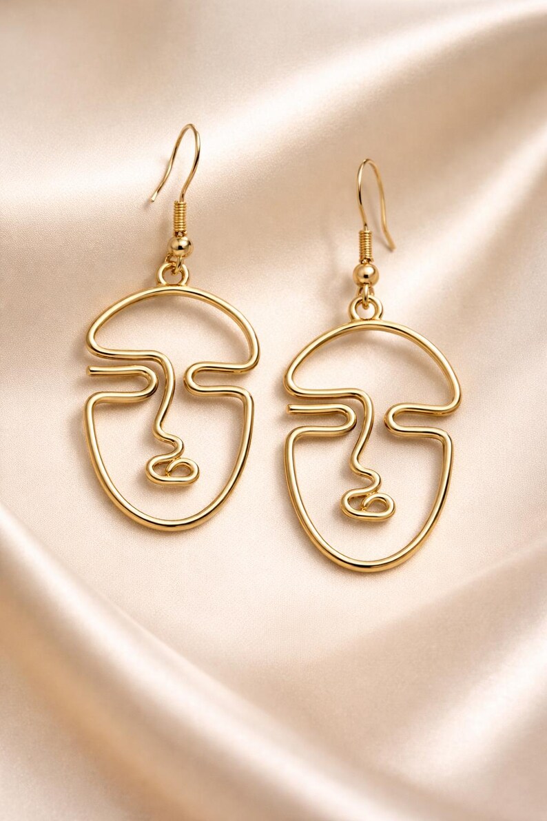Boucles d’oreilles visage abstrait en fil doré, design minimaliste fait main. Lignes continues formant un visage artistique, finition dorée douce type champagne. Présentées sur tissu beige clair avec plis élégants, lumière naturelle douce et ombres légères, style raffiné et moderne type bijouterie parisienne.