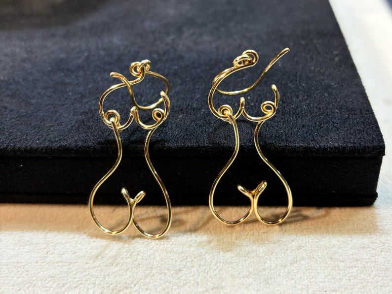 Boucles d’oreilles en fil doré représentant un corps féminin stylisé, accrochées sur un support noir.