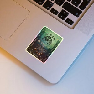 May include: A rectangular sticker with a holographic border, featuring the text "BE WARY, BE CLEVER, BE GOOD" around a tree illustration. The sticker has a green and gold color scheme and is placed on a laptop.
